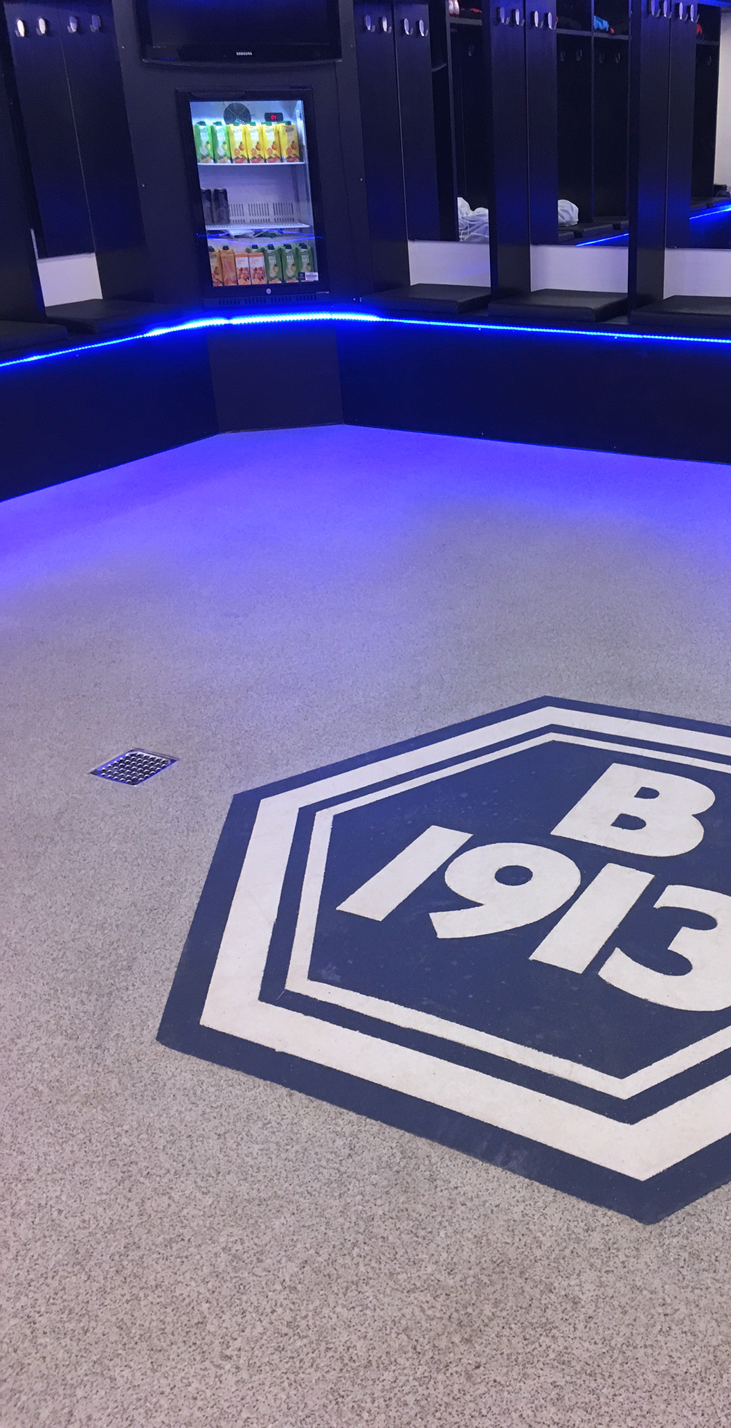 Blå vådrumsgulv med B1913 logo på, skridsikre gulve og fugefri gulve i bad- og omklædningsrum hos fodboldklubben B1913 med blå led lys langs kanten og glaskøleskab med drikkevarer leveret af ekspert i materialer til gulve Stonewalk